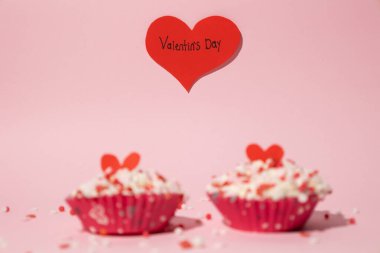 Two out of focus cupcakes decorated with colorful candies. Behind them there is a red heart with a written message that says Valentine's Day. On a pink background. Concept of love and affection. clipart