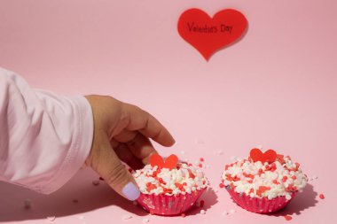 Female hand placing cupcakes decorated with colorful candies on a pink background. Valentine's Day. Love and affection concept clipart