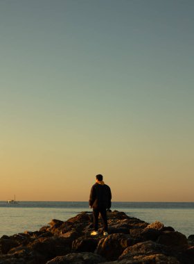 Bir kış sabahı kayaların ortasında durup denize bakan erkek fotoğrafçının dikey görüntüsü. Elinde bir kamera tutuyor ve sımsıkı sarılıyor. Yaşam biçimi