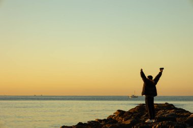 Neşeli fotoğrafçı bir kış sabahı elinde bir kamerayla denize bakarken taşların ortasında elini kaldırıyor. Yaşam biçimi