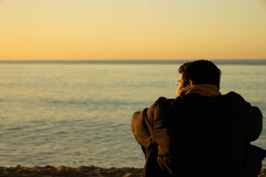 Genç adam sahilde oturmuş, altın bir kış sabahı ufka bakıyor. Yaşam biçimi