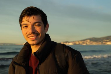 Portrait of cheerful freckled young man. with morning sun falling on his face with blue sea in background. lifestyle clipart