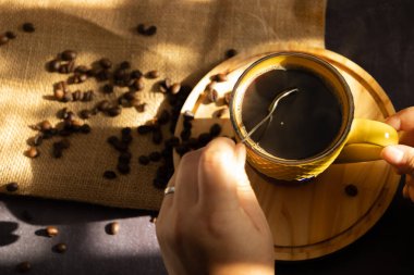 Female hand stirring hot coffee in a yellow cup with a spoon and coffee beans scattered on the bottom in the morning clipart