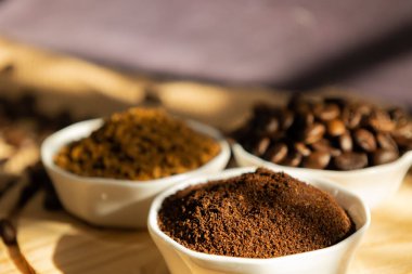 Close up of small porcelain bowls with different types of coffee. beans, ground and instant, on a wooden plate on a dark background with sunlight coming through the window and scattered coffee beans clipart