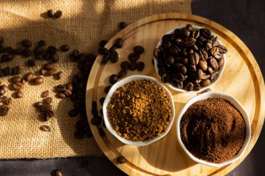 Küçük porselen kaselerin farklı kahve çeşitleriyle üst görünümü. Fasulye, zemin ve hazır. Koyu bir zemin üzerinde, ahşap bir tabakta. Pencereden güneş ışığı ve dağınık kahve çekirdekleri geliyor.