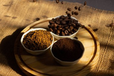 Kahve çekirdeklerinin farklı kahve çeşitleriyle küçük porselen kaselere düşüşünün üst görüntüsü. Fasulye, zemin ve hazır, tahtadan bir tabakta, karanlık bir arka planda. Pencereden güneş ışığı geliyor.