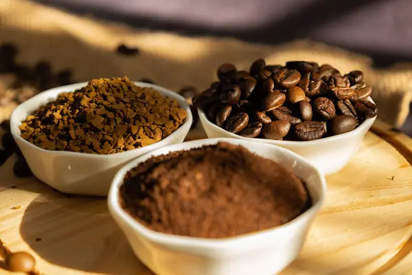 Hazır kahve ve kahve çekirdekleri beyaz porselen kaselerde, ahşap bir tabakta ve koyu bir arka planda dağınık kahve çekirdekleri sabah güneşi tarafından aydınlatılmış.