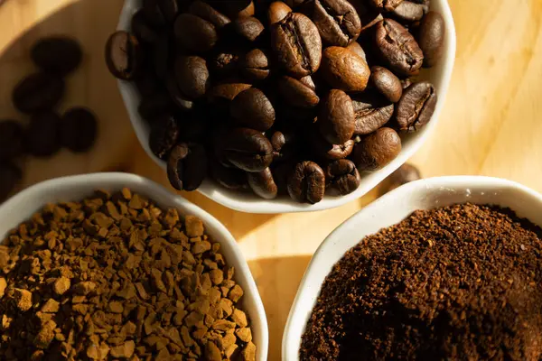 Farklı türde kahvenin olduğu küçük porselen kaselere aşırı yakın çekim. Fasulye, zemin ve hazır. Koyu bir zemin üzerinde, ahşap bir tabakta. Pencereden güneş ışığı ve dağınık kahve çekirdekleri geliyor.