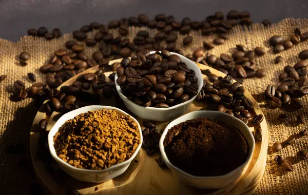 Kahve çekirdekleri farklı kahve çeşitleriyle küçük porselen kaselerin yanına saçılmış. Fasulye, zemin ve hazır, tahtadan bir tabakta, karanlık bir arka planda. Pencereden güneş ışığı geliyor.