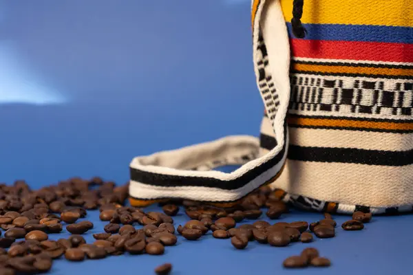 Kahve çekirdeklerini yakın plan koyu mavi arkaplana serpiştirilmiş halde sabah güneş ışınlarıyla aydınlatılmış tipik bir Kolombiya çantasının yanında. Kahve ve Kolombiya kültürü kavramı