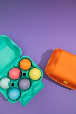 Vertical image in top view of colorful eggs in a blue painted basket next to an orange one on a purple background. Easter painted eggs clipart