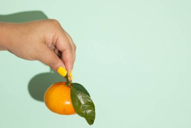 Açık yeşil izole bir zemin üzerinde parmaklarıyla bir portakalı tutan dişi el, C vitamini bakımından zengin sağlıklı tropikal meyve.