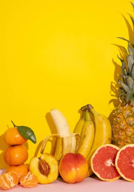 Sarı zemin üzerinde pembe, ananas, muz, şeftali, mandalina ve greyfurt bulunan sıcak renkli tropikal meyvelerin dikey görüntüsü. Tek renkli sarı sağlıklı yiyecekler.
