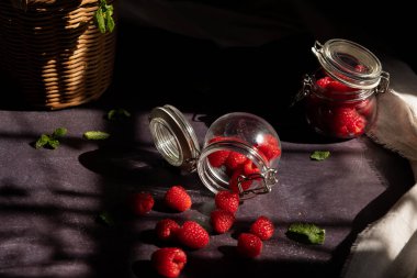 Koyu bir zemin üzerinde uzanan cam kavanozların üst görüntüsü etrafa saçılmış ahududu ve etrafta nane yaprakları var ve pencereden güneş geliyor. Kara meyve yemeği.