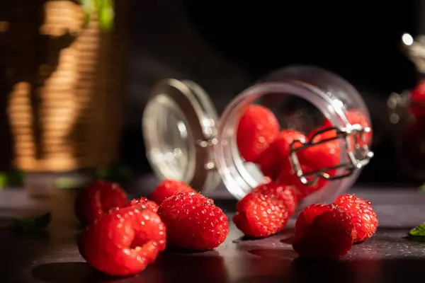 Koyu bir zemin üzerinde uzanan cam kavanozları kapatın etrafa saçılmış ahududu ve etrafa nane yaprakları saçın ve pencereden güneş girsin. Kara meyve yemeği.