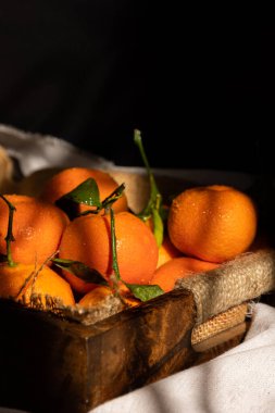 Sabah güneşi tarafından pencereden aydınlatılan, rustik ahşap bir tepside taze toplanmış portakalların dikey görüntüsü. Kara yemek.