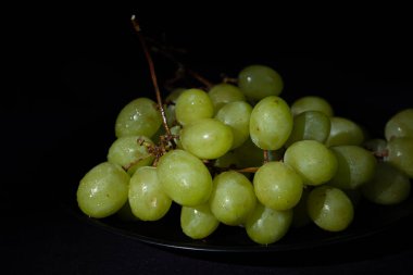 Arka planda siyah bir tabakta bir demet yeşil üzüm. Yuvarlak ve sulu meyve