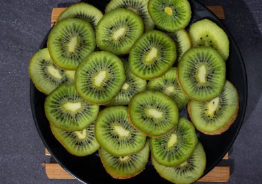 Siyah bir tabakta koyu bir arka planda kesilmiş taze kivilerin üst görüntüsü. Kıllı bir deriyle kaplı elips şeklinde yeşilimsi yiyecekler. Koyu yiyecek