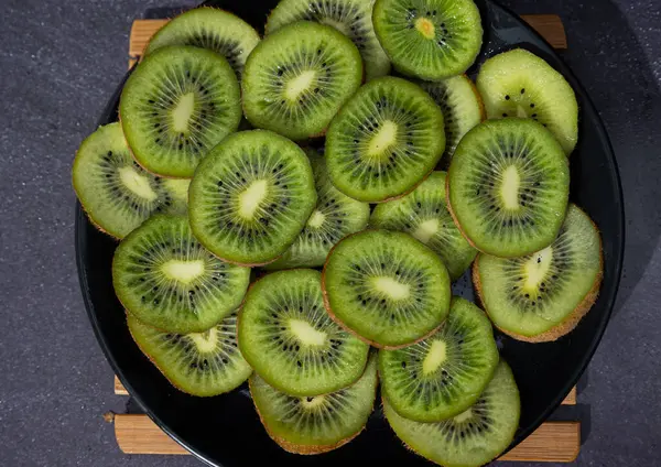 Siyah bir tabakta koyu bir arka planda kesilmiş taze kivilerin üst görüntüsü. Kıllı bir deriyle kaplı elips şeklinde yeşilimsi yiyecekler. Koyu yiyecek