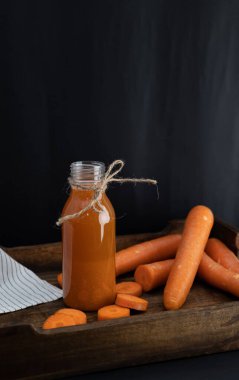Koyu bir arka planda, ahşap bir tepsiye saçılmış havuçların yanında şişedeki havuç püresinin dikey görüntüsü.