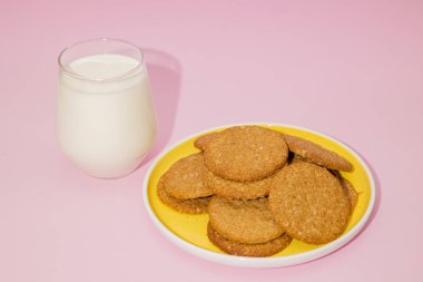 Sarı bir tabakta kepekli yulaf ezmeli kurabiyeler yanında bir bardak süt pastel pembe arka planda, buğday unuyla yulaf gevreğiyle karıştırılmış kurabiyeler, geleneksel pişirme.
