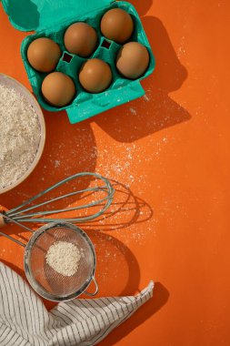Turuncu arka planda kurabiye yapmak için yumurta, un ve mutfak aletlerinin dikey görüntüsü