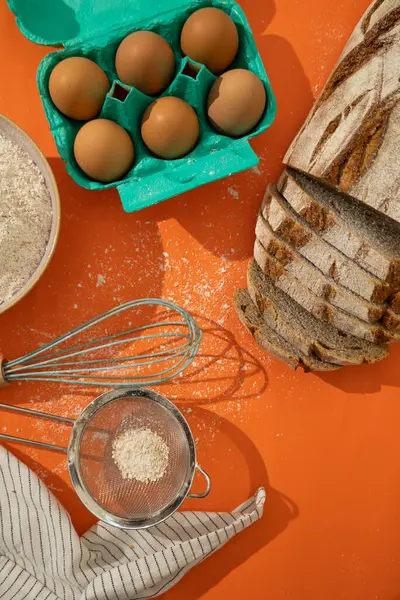 Buğday ekmeğinin üst görüntüsü un tozlu ve turuncu arka planda dilimlenmiş ve yapımında kullanılan malzemelerle birlikte. Yüksek lifli besleyici yiyecekler.