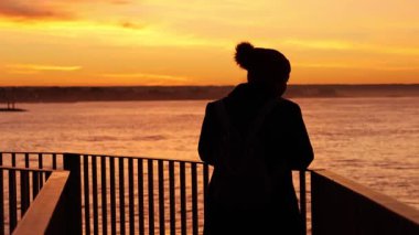 Genç bir kadın kışın sarılıp deniz kıyısındaki bir iskelede altın gün doğumunu seyrediyor. Açık hava yaşam tarzı