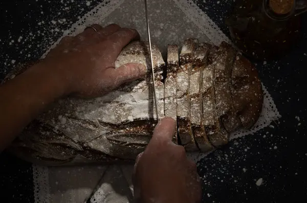 Erkek el, koyu bir arka planda, un tozuyla kaplanmış bir buğday ekmeğini dilimliyor. Yüksek lif içerikli besleyici yiyecekler, koyu besinler.
