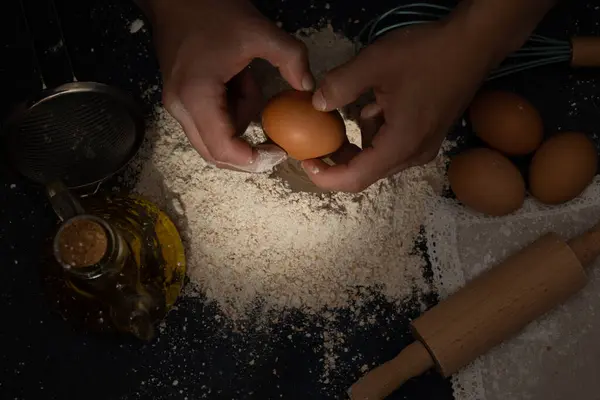 Erkek eller tam buğday ununa yumurta kırıyor. Ekmek yapmak için malzemeler ve malzemeler koyu bir arka planda. Kara yemek.