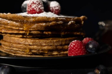 Üzerine kırmızı ahududu ve yaban mersini serpiştirilmiş krep ve koyu arkaplanda siyah bir tabakta krema şekeri. Ev yapımı yemekler, karanlık yemekler.