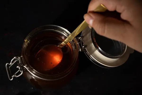 Kadın elinin elinde altın kaşıkla koyu bir arka plandaki cam bir kutudan bal çıkardığı güzel ve yapışkan bir sıvının tatlı bir tadı olduğu bir görüntü.