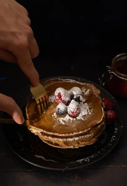 Siyah bir plaka üzerinde pudra şekeri, kırmızı ahududu ve yaban mersiniyle istiflenmiş krepleri kesmek için çatal bıçak takımı kullanan erkeklerin dikey görüntüsü. Ev yapımı yemekler, karanlık yemekler.