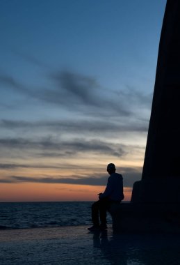 Bir yapının üzerine oturmuş kışın deniz kenarında gün batımını izleyen bir adamın silueti.