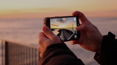 Deniz kıyısındaki bir iskelede altın gündoğumunu kaydetmek için cep telefonunu kullanan bir adamın ellerini kapatın. Açık hava yaşam tarzı