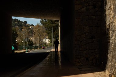 Güneşli bir günde, bir kasabanın varoşlarında yol tünelinden geçen bir kadının silüeti.