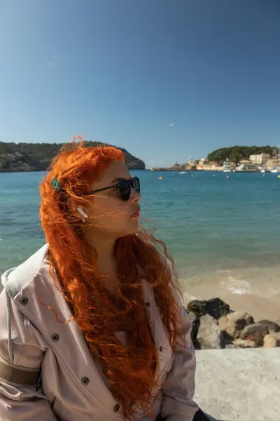 Çilli, kızıl saçlı, gözlüklü bir kadının, güneşli bir günde arka planda mavi deniz olan manzaraya bakarken deniz kenarında oturup müzik dinlediği dikey görüntüsünü kapat.