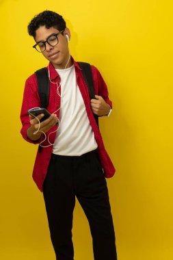 Genç kahverengi tenli Latin bir adamın dikey görüntüsü, günlük kırmızı kıyafetler giyen bir üniversite öğrencisi, sarı arka planda mutlu bir şekilde fotokopi alanı olan akıllı telefonunu kullanıyor.