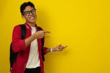 Genç, gülümseyen, kahverengi tenli, kırmızı günlük kıyafetler giyen Latin öğrenci ellerini sarı arkaplanda yazı için bir yeri işaret ediyor. Reklam için telif alanı olan grafik bir kaynak.