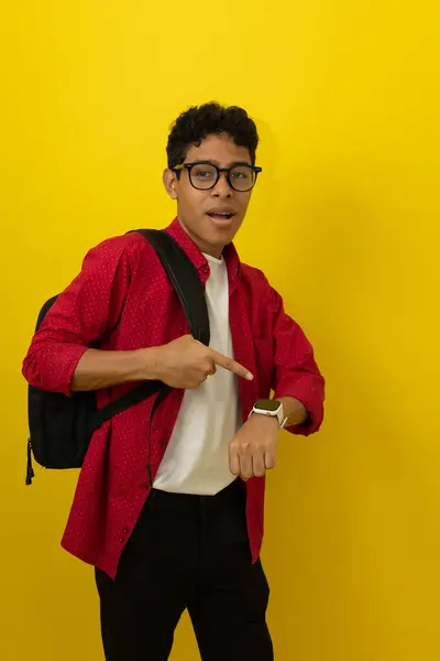 Kahverengi tenli, gözlüklü ve çantalı genç bir Latin öğrenci saati işaret ederek sarı arka planda fotokopi alanı olan saati gösteriyor.