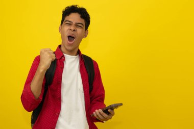 Genç Latin bir adam, elinde bir cep telefonuyla fotokopi çekilen sarı bir arkaplanda kutlama jesti yapıyor.