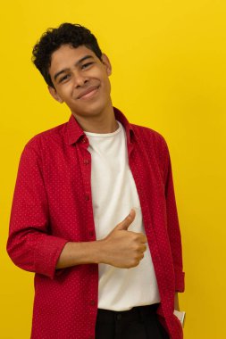 Genç, kahverengi tenli Latin bir adamın dikey görüntüsü gündelik kırmızı elbiseler giymiş, başparmağıyla sarı arka planda fotokopi alanı olan.