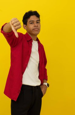 Genç, kahverengi tenli Latin bir adamın dikey görüntüsü. Günlük kırmızı kıyafetler giyiyor. Başparmakları aşağıda, sarı arka planda fotokopi alanı var.