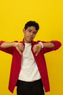 Genç, kahverengi tenli Latin bir adamın dikey görüntüsü. Günlük kırmızı giysiler giyiyor. İki başparmağı da aşağıda, sarı arka planda fotokopi alanı var.