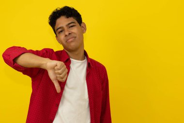 Genç kahverengi tenli Latin adam, günlük kırmızı kıyafetler giyiyor. Başparmakları aşağıda, sarı arka planda fotokopi alanı var.