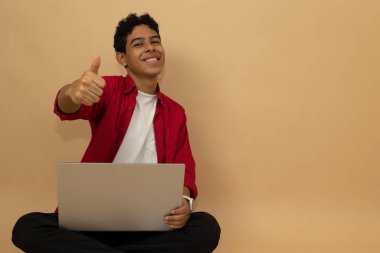 Kırmızı ve siyah gündelik kıyafetler giyen neşeli Latin bir adam oturmuş dizüstü bilgisayarını açık kahverengi bir arka planda reklam alanında kullanırken bir jest yapıyor.