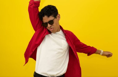 Neşeli, kahverengi tenli, sıradan kırmızı elbiseler giyen Latin adam, fotokopi çekilen sarı arka planda siyah gözlüklerle dans ederken ellerini kaldırıyor.