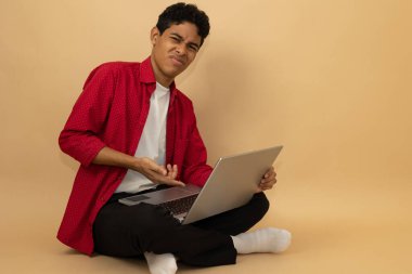 Kırmızı ve siyah gündelik giysiler giyen Latin kökenli genç bir adamın profil görüntüsü. Açık kahverengi arka planda, reklam alanı olan dizüstü bilgisayarını kullanarak oturuyor ve parmağıyla işaret ediyor.