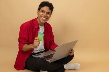 Gülümseyen kahverengi tenli Latin bir adam elinde bir kartla oturmuş açık kahverengi bir arka planda reklam alanı olan bilgisayarından alışveriş yapıyor.