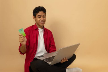 Kahverengi tenli neşeli Latin bir adam elinde bir kartla oturmuş açık kahverengi arka planda reklam alanı olan bilgisayar alışverişine bakıyor.